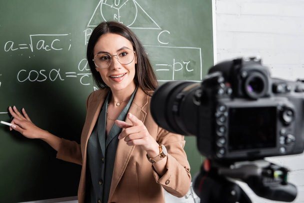 Smiling teacher holding chalk near chalkboard with formulas and digital camera  - 写真・画像