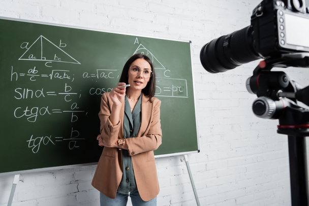 Δάσκαλος κρατώντας κιμωλία και μιλώντας κοντά σε μαυροπίνακα με τύπους και ψηφιακή φωτογραφική μηχανή  - Φωτογραφία, εικόνα