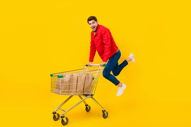 Full length body size view of attractive funny cheerful guy holding cart jumping isolated over bright yellow color background. - Foto, imagen
