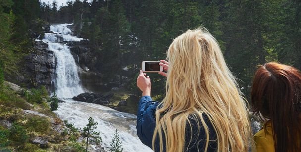 Žena fotografovat vodopád na mobilu během túry v přírodě s kamarádem.Dvě ženy na túře v přírodě společně fotit vodopád pomocí mobilního telefonu - Fotografie, Obrázek