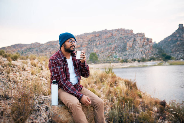 A young adult man takes a break from hiking drinks coffee by the lake view. High quality photo - Photo, Image