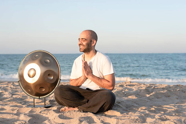 ένας χαμογελαστός μουσικός γιόγκι διαλογίζεται πριν παίξει στο χερούλι ένα πνευματικό όργανο που κάθεται στην παραλία - Φωτογραφία, εικόνα