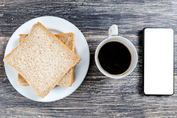 Sliced bread on the plate and cup of coffee with a mobile phone with a blank screen for copy space on wooden background - Foto, Imagen