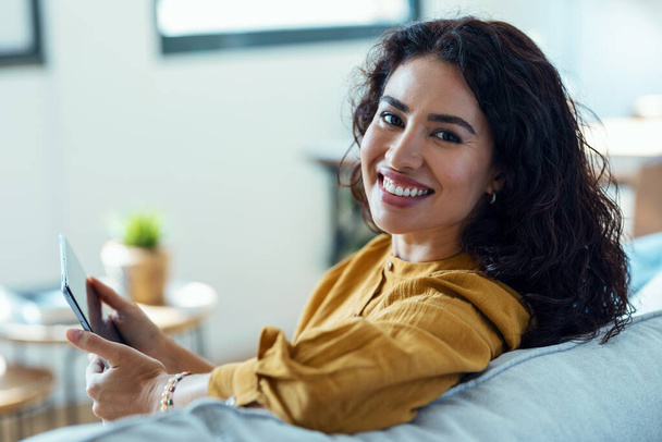 Φωτογραφία της όμορφης νεαρής γυναίκας που χρησιμοποιεί το ψηφιακό tablet της για ανάγνωση, ενώ κάθεται στον καναπέ στο σπίτι. - Φωτογραφία, εικόνα
