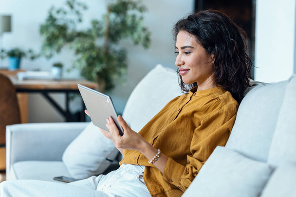 Foto de una mujer joven y bonita usando su tableta digital para leer mientras está sentada en el sofá en casa
. - Foto, imagen