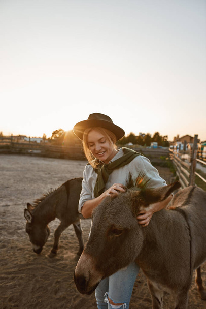 Caucasian woman caressing cute donkey in paddock in village or ranch. Young smiling blonde woman. Modern hipster farm lifestyle. Agriculture and farming. Countryside landscape. Sunset time - Photo, image