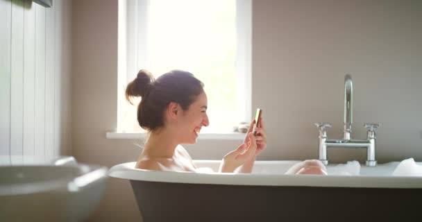 Une femme heureuse souffle un baiser lors d'un appel vidéo sur son téléphone portable dans un bain moussant. - Séquence, vidéo