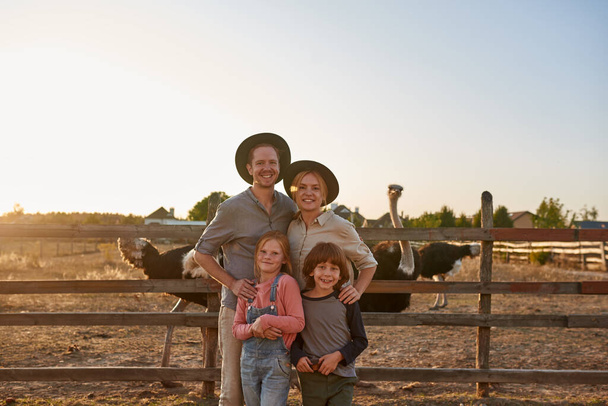 Young happy caucasian family near paddock with ostriches in village or ranch. Modern farm lifestyle. Mother, father, son and daughter looking at camera. Agriculture. Countryside landscape. Sunset time - Foto, immagini