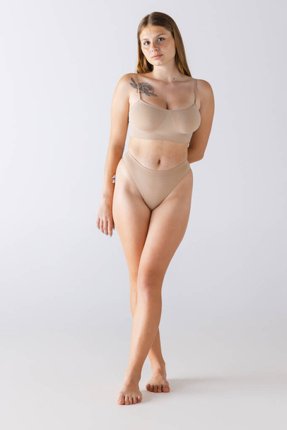 Portrait of young tender girl posing in beige underwear isolated over grey studio background. Concept of beauty, body and skin care, spanx inner wear, shapewear, health, plastic surgery, cosmetics, ad - Φωτογραφία, εικόνα