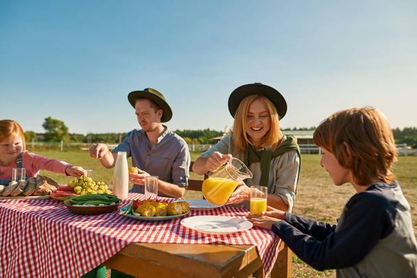 Young caucasian hipster farm family having lunch with fresh organic products in countryside outdoors. Mother pouring juice for son. Father and daughter eating fruits. Modern farm lifestyle. Sunny day - Φωτογραφία, εικόνα