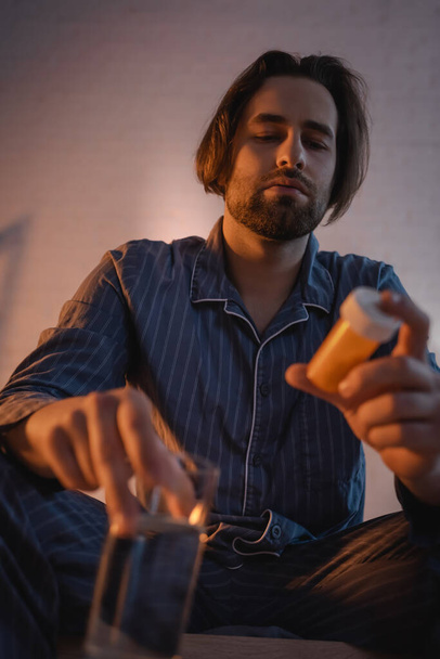 Low angle view of man with insomnia holding blurred water and pills in bedroom  - Foto, afbeelding