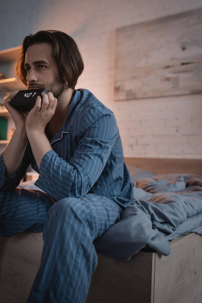 Brunette man in pajama holding alarm clock on bed at night  - Fotografie, Obrázek