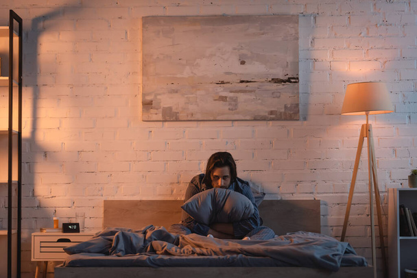 Tired man holding pillow and looking at camera on bed at night  - Valokuva, kuva