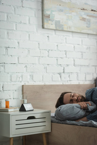 Молодой человек с закрытыми глазами лежит на подушке возле таблеток и будильника в спальне  - Фото, изображение