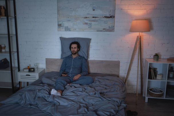 Uykusuzluk çeken bir adam gece yatakta otururken uzaktan kumanda tutuyor.  - Fotoğraf, Görsel