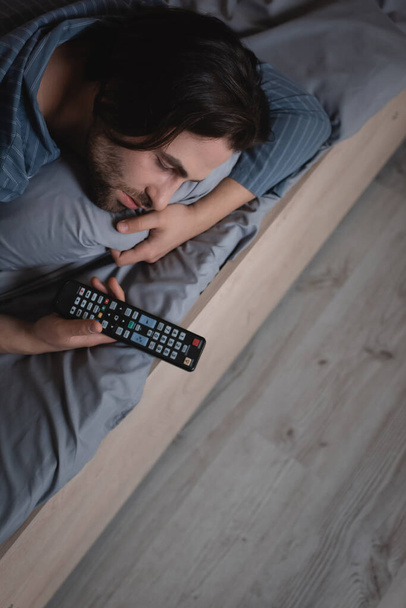 Vysoký úhel pohledu na muže držícího dálkové ovládání, když spí v noci na posteli  - Fotografie, Obrázek