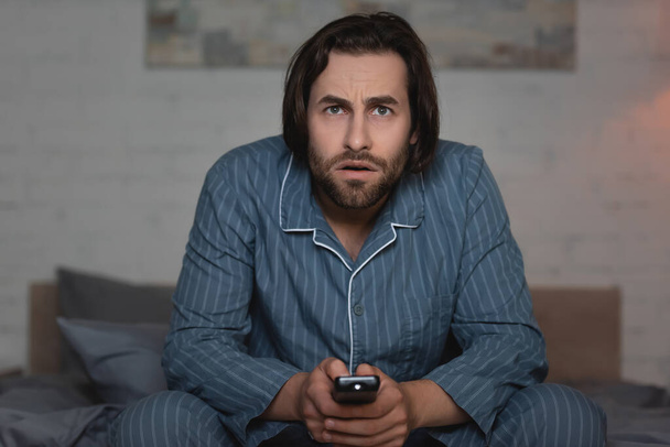 Man in pajama holding remote controller while watching tv on blurred bed  - Foto, afbeelding