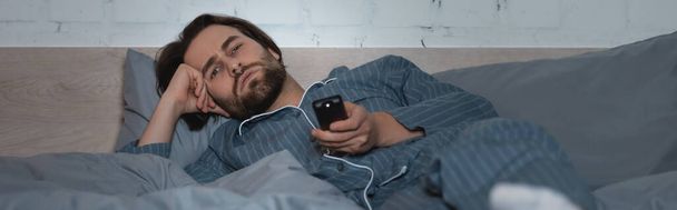 Εξαντλημένος άνθρωπος βλέποντας τηλεόραση, ενώ πάσχουν από αϋπνία στο κρεβάτι, πανό  - Φωτογραφία, εικόνα