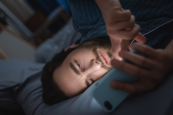 Bearded man with insomnia using mobile phone on bed  - Фото, изображение