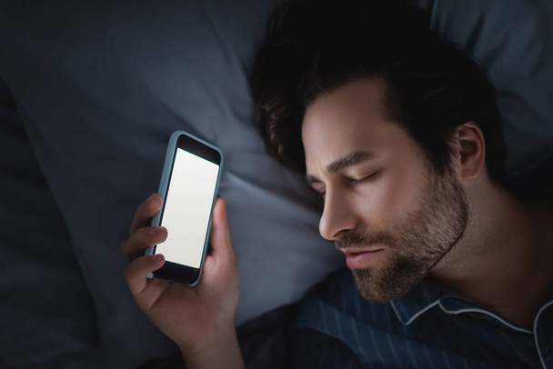 Top view of man in pajama holding smartphone with blank screen while sleeping on bed  - Foto, Bild