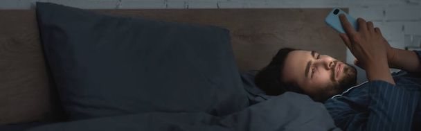 Бородатий чоловік, який страждає від безсоння і використовує смартфон на ліжку вдома, банер
  - Фото, зображення
