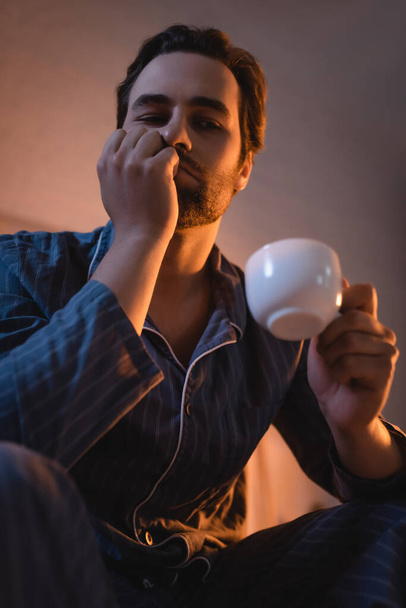 Low angle view of sad man with insomnia holding cup of coffee at home  - Foto, Bild