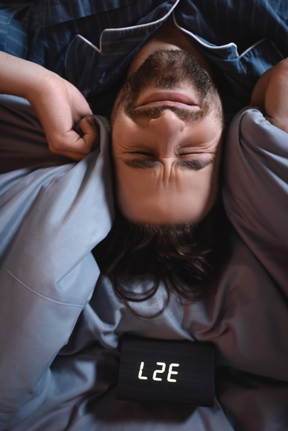 Top view of tensed man covering ears near alarm clock on bed at night  - Fotó, kép