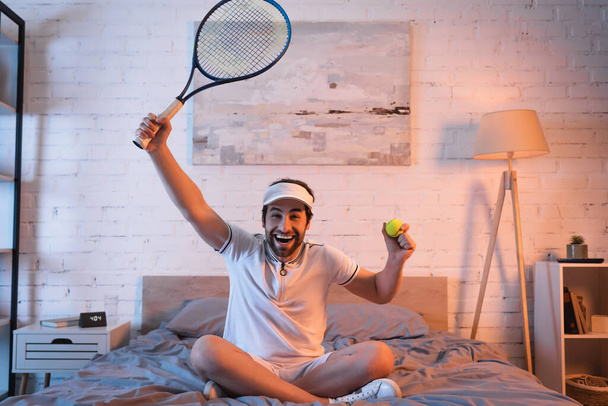 Cheerful sleepwalker in sportswear holding tennis rocket and ball on bed  - 写真・画像