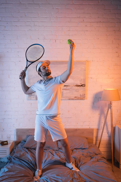 Somnambulist holding tennis ball and rocket while standing on bed at home  - Valokuva, kuva