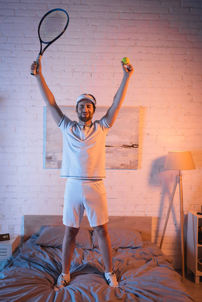 Excited somnambulist holding tennis ball and rocket on bed at night  - Foto, Imagem