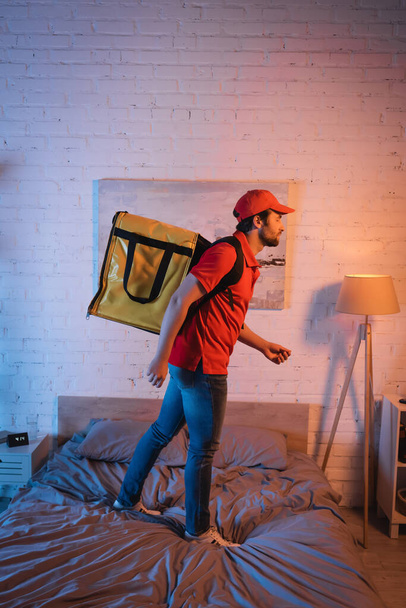 Side view of sleepwalker in uniform with thermo backpack standing on bed at night  - Fotoğraf, Görsel