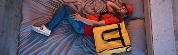 Teslimat üniformalı genç bir uyurgezer yatakta termo sırt çantasının yanında uyuyor.  - Fotoğraf, Görsel