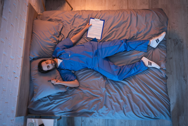 Top view of sleepwalker in doctor uniform holding clipboard and pen while sleeping on bed  - Foto, imagen
