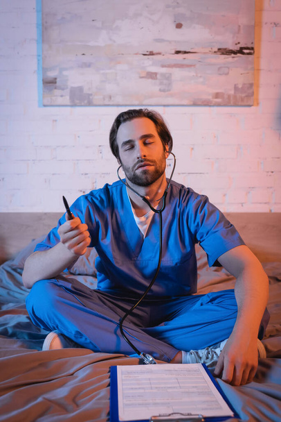 Young sleepwalker in doctor uniform and stethoscope holding pen near clipboard on bed  - Photo, Image