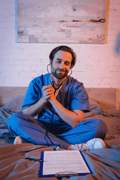 Smiling somnambulist in doctor uniform and stethoscope sitting near clipboard on bed  - 写真・画像
