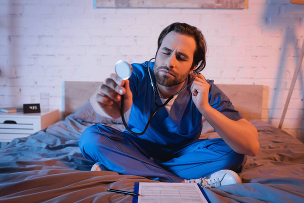 Sleepwalker in doctor uniform holding stethoscope near clipboard on bed at night  - Фото, изображение