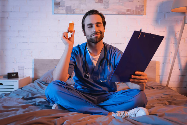 Smiling sleepwalker in doctor uniform holding clipboard and pills on bed at night  - Foto, imagen