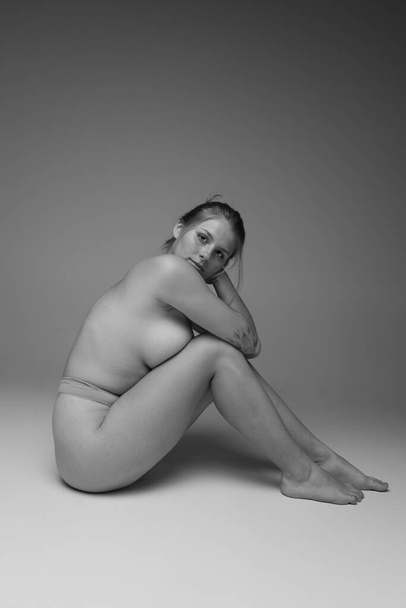 Portret młodej atrakcyjnej kobiety siedzącej na podłodze, pozującej w bieliźnie. Czarno-białe zdjęcia. Pozytywność ciała. Pojęcie piękna, pielęgnacji ciała i skóry, zdrowia, chirurgii plastycznej, kosmetyków, reklamy - Zdjęcie, obraz