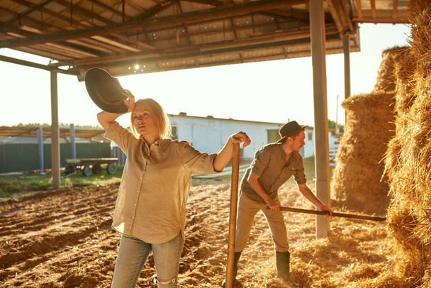 Man collecting hay in bale with pitchfork while his tired girlfriend wiping forehead during having break at work. Young caucasian couple. Modern hipster farm family lifestyle. Agriculture. Sunny day - Fotoğraf, Görsel
