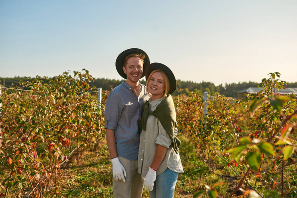 Front view of young european couple hugging and looking at camera in countryside garden. Man and girl relationship and spend time together. Modern hipster farm family lifestyle. Agriculture. Sunny day - 写真・画像