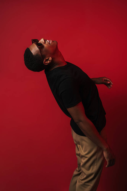 Young black man wearing sunglasses screaming and looking upward isolated over red wall - Foto, Bild