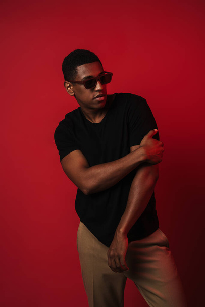 Young black man wearing sunglasses posing and looking aside isolated over red wall - Fotó, kép