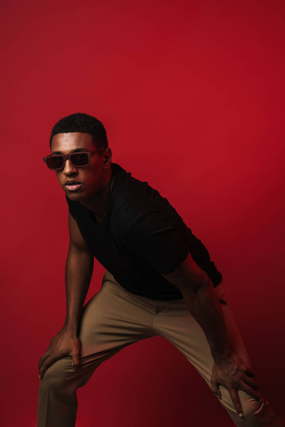 Young black man wearing sunglasses posing and looking at camera isolated over red wall - Foto, Bild