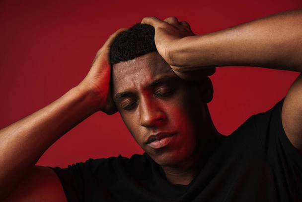 Joven hombre negro con dolor de cabeza sosteniendo la cabeza mientras posa en la cámara aislado sobre la pared roja - Foto, Imagen