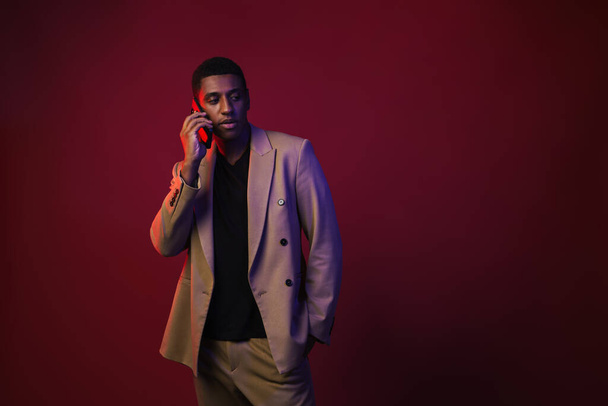 Young black man dressed in jacket talking on mobile phone isolated over red wall - Fotó, kép