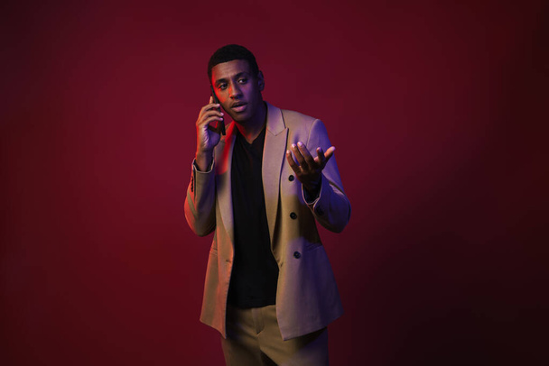 Young black man dressed in jacket gesturing while talking on cellphone isolated over red wall - Фото, изображение