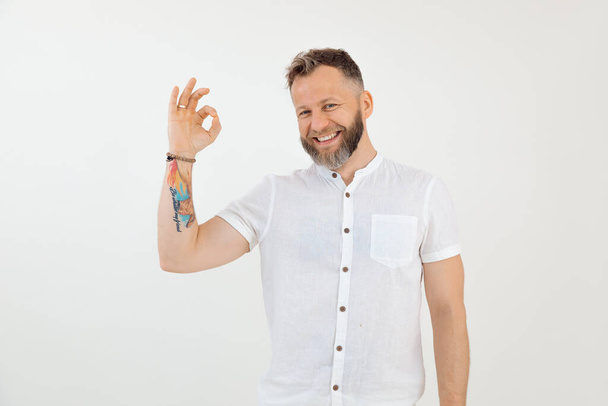 Bearded smiling tattooed businessman looking at camera in white outfit showing ok sign, presenting success in career. Advertisement for massage, physiotherapy treatment. White isolated background. - 写真・画像