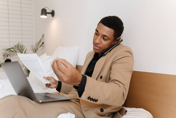 Black man talking on cellphone while working with laptop and documents on bad in hotel - Photo, image