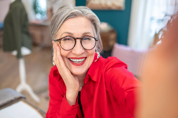 Good moments. Happy stylish gray-haired woman in glasses touching face with palm smiling at camera sitting in her atelier - 写真・画像