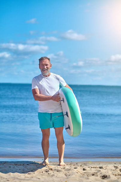 Great day. Adult grey-haired bearded man holding surfboard smiling at camera standing with his back to sea on sunny day - Fotografie, Obrázek